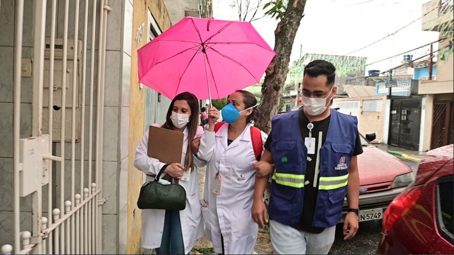 Na imagem, há três pessoas andando na calçada de uma rua, sendo duas mulheres e um homem. As duas mulheres estão usando jaleco e seguram um guarda-chuva rosa. Ao lado delas, um homem usa colete azul.